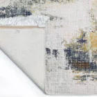 A close-up of a rug's corner, with a white, gold, and dark blue pattern, with a white fringe at the edge, from Qaleen Carpets