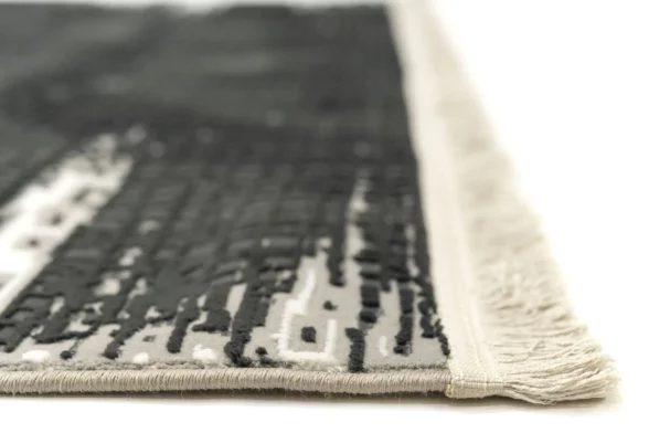 A close-up of a rug with a fringe border, made by Qaleen Carpets, featuring a pattern of dark and light grey, with a white