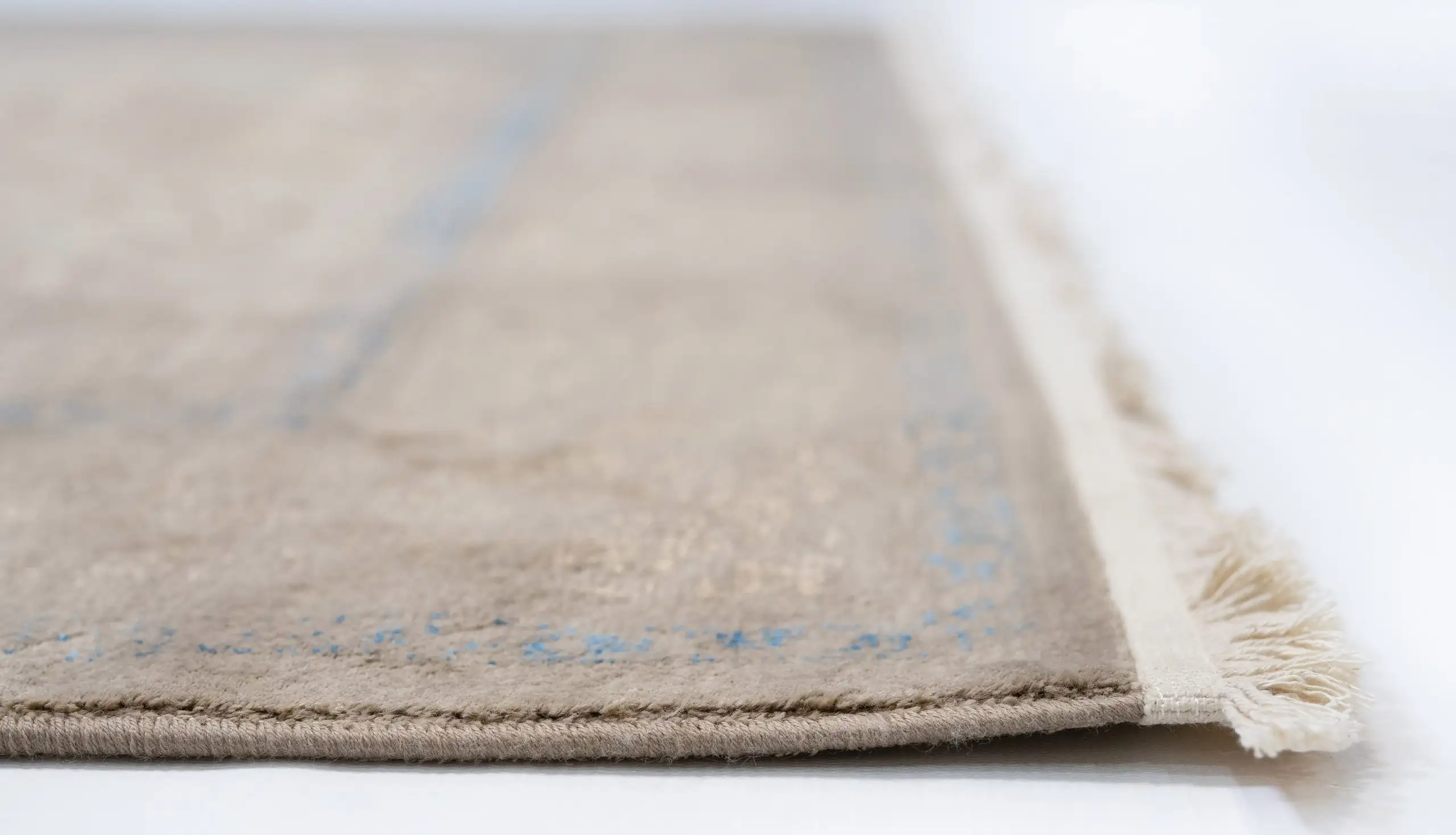 A close-up of a beige rug with a subtle blue pattern, with a fringe on the right side, made by Qaleen Carpets