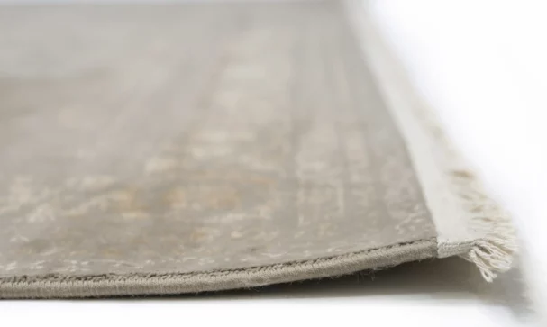 A close-up of a Qaleen Carpets rug with a soft, neutral color and subtle pattern, featuring a fringe on one edge
