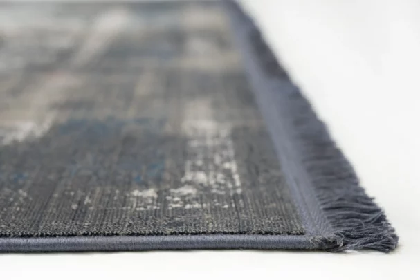 A close-up of a gray and blue rug with a fringe on one side, made by Qaleen Carpets