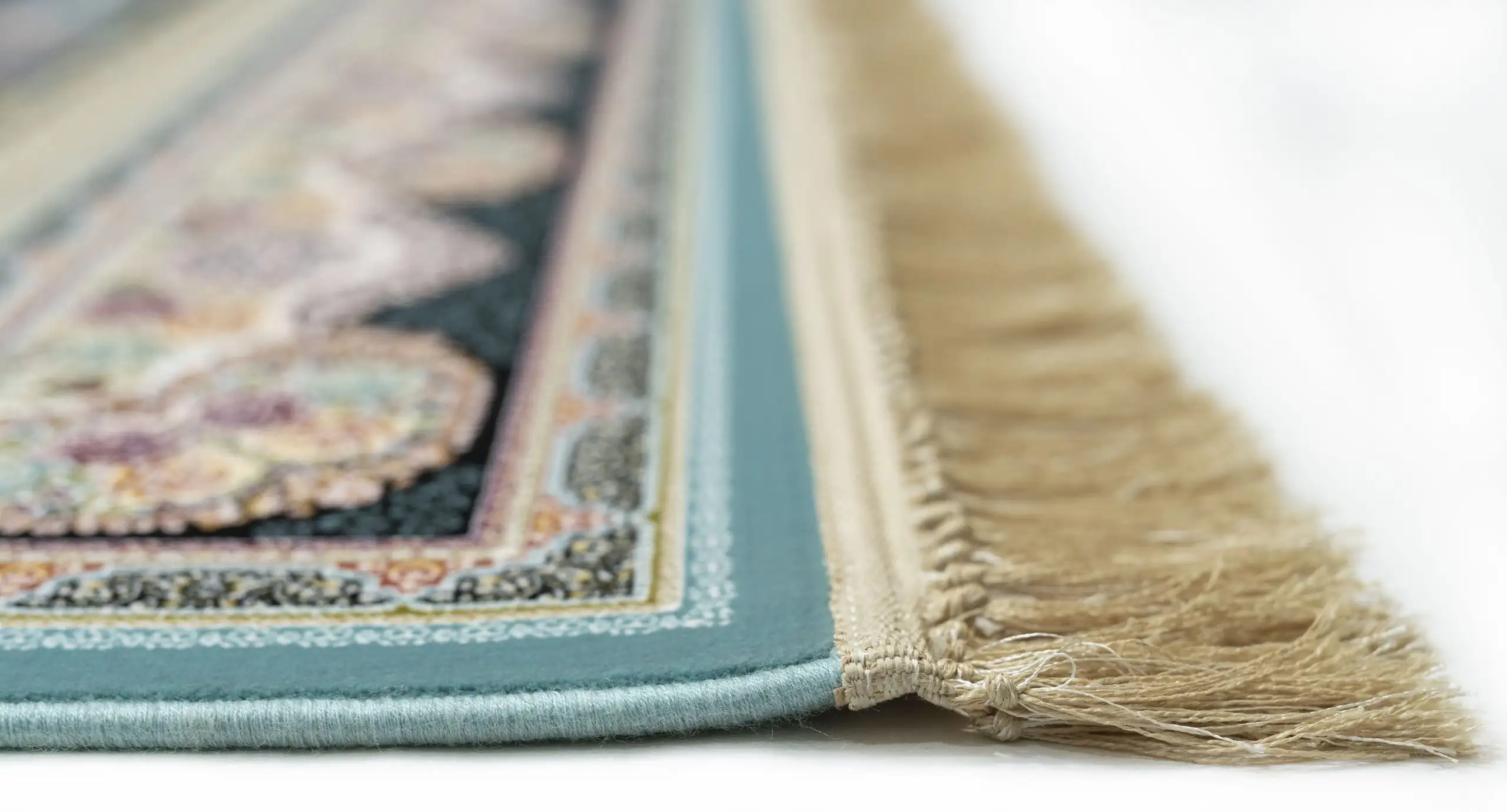 A close-up of a rug with a blue border and a patterned design, with a fringe at the bottom, made by Qaleen Carpets