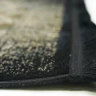 A close-up of a black and gold patterned rug with a fringe on one edge, made by Qaleen Carpets