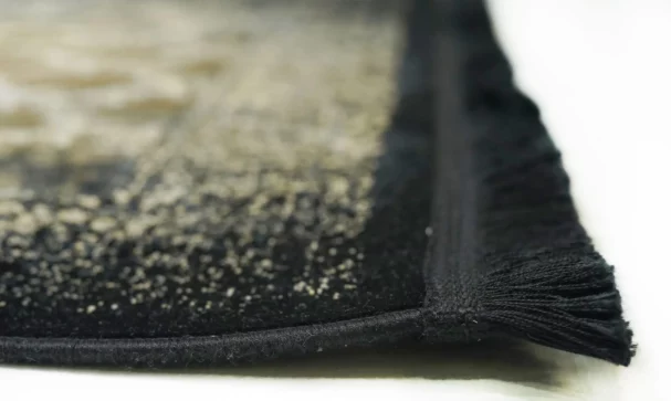 A close-up of a black and gold patterned rug with a fringe on one edge, made by Qaleen Carpets