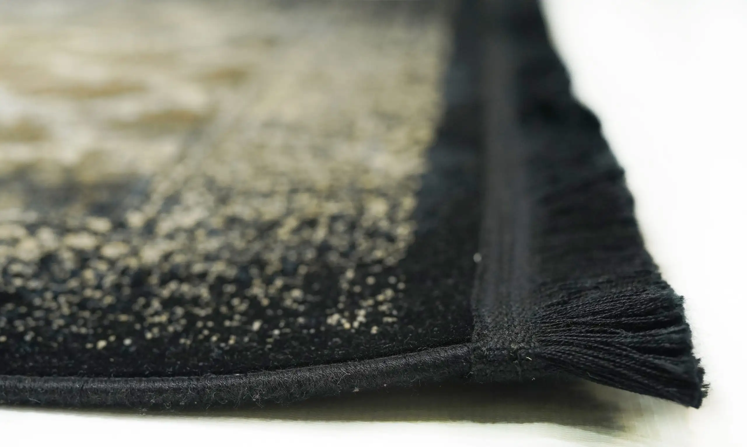 A close-up of a black and gold patterned rug with a fringe on one edge, made by Qaleen Carpets