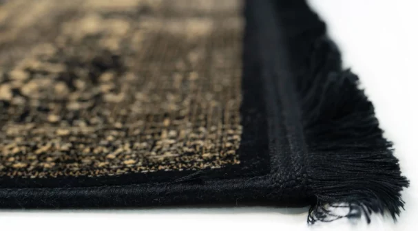 A close-up of a Qaleen Carpets rug with a black and gold pattern, featuring a fringe on one edge