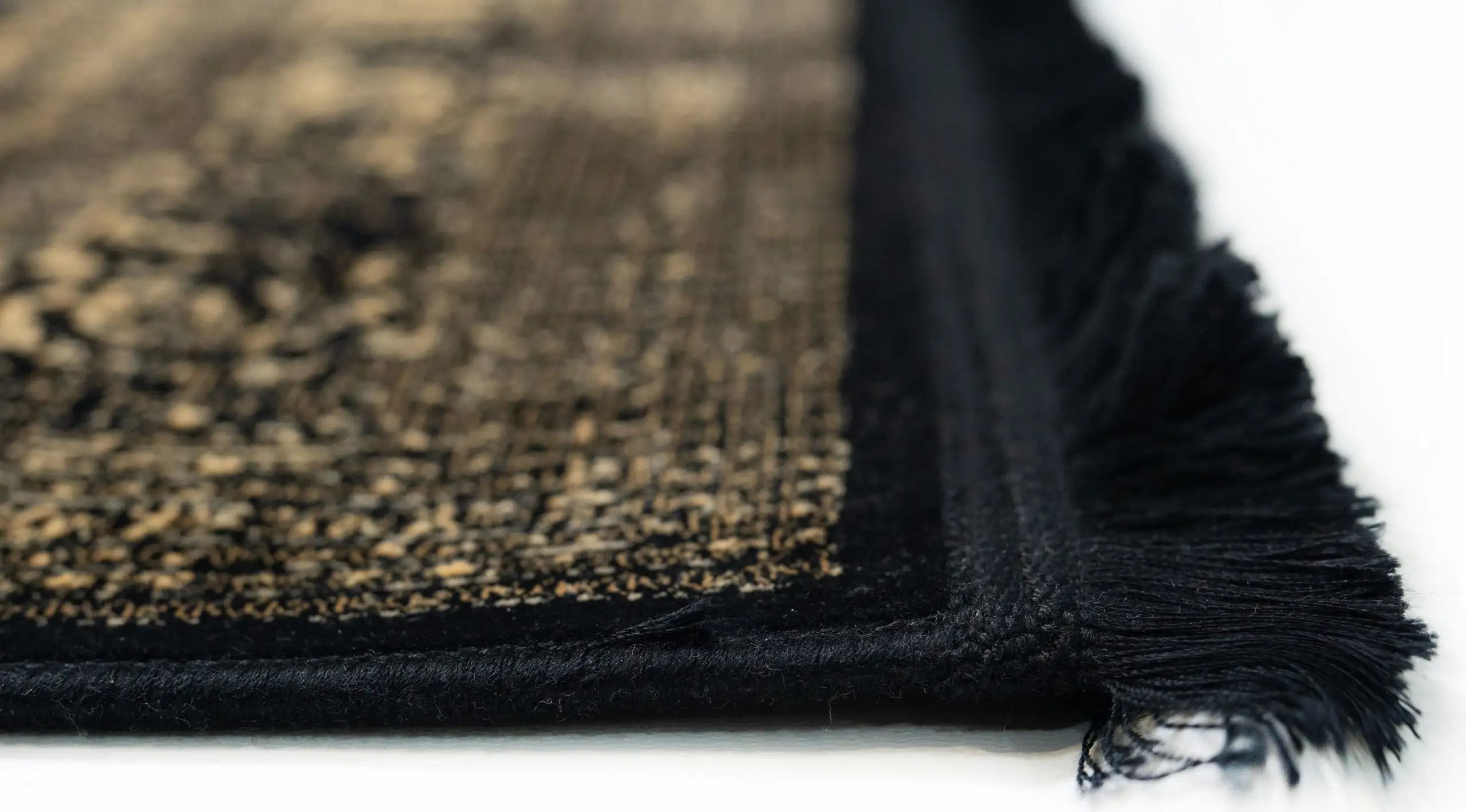 A close-up of a Qaleen Carpets rug with a black and gold pattern, featuring a fringe on one edge