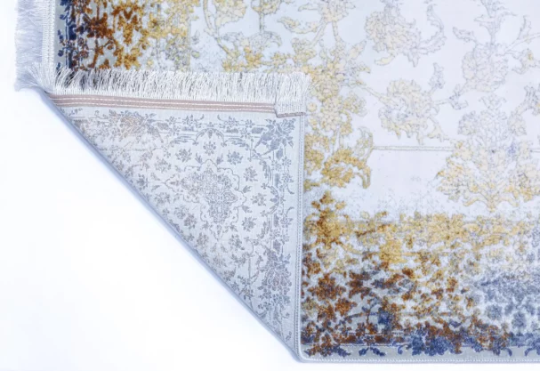 A corner of a Qaleen Carpets rug with a white background, featuring a mix of gold, blue, and brown patterns, with a fringe